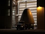 Goddard, the first development vehicle in the New Shepard program, is transported out of a large warehouse on a truck.