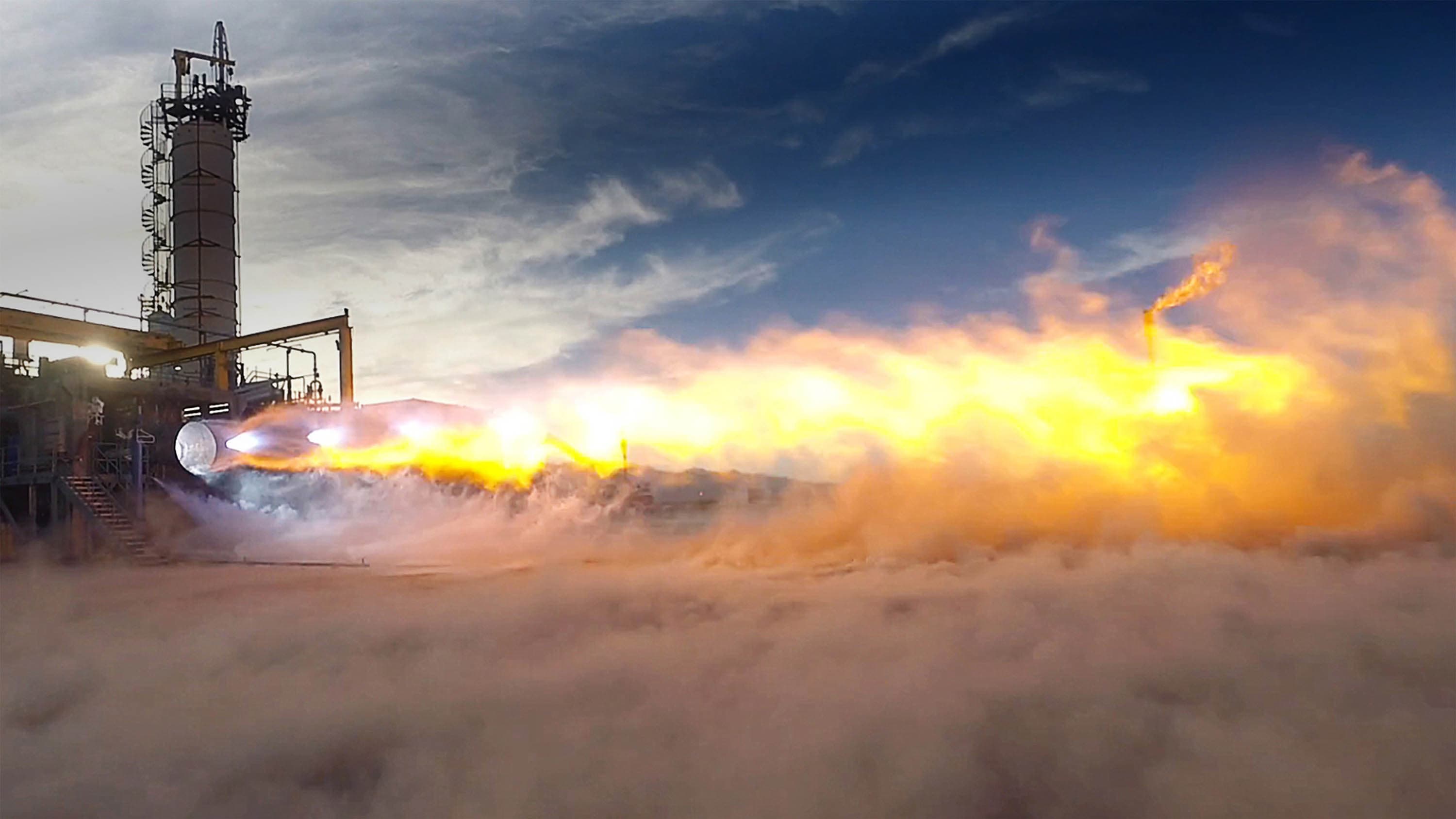 An engine is mounted on a large metal structure expelling a horizontal plume of fiery exhaust with mach diamonds. White smoke and dust fills the bottom half of the frame.