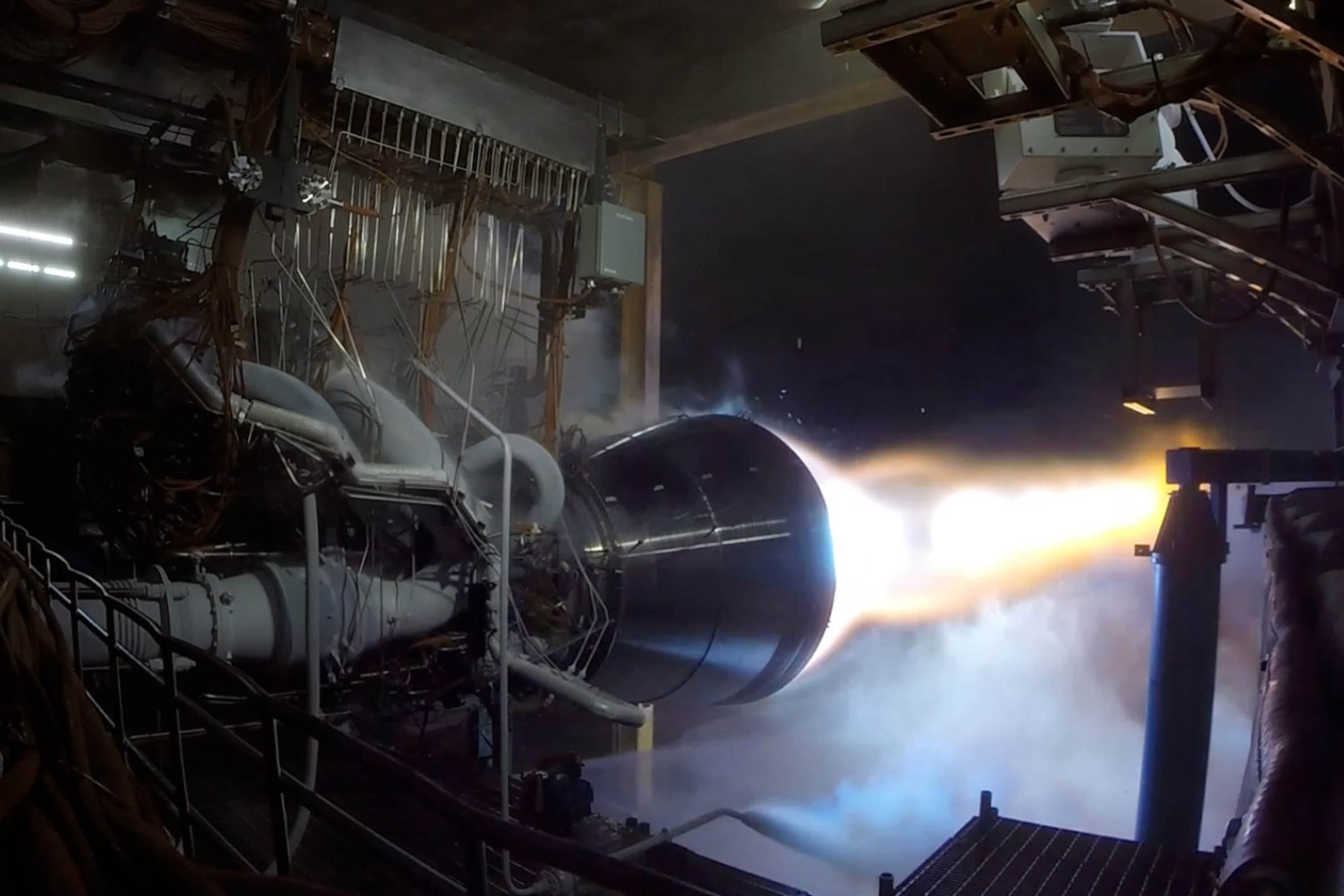 A view from inside the test stand of the engine affixed horizontally to the metal structure. A fierce plume of white and orange exhaust is expelled from the black nozzle in the center of the frame.