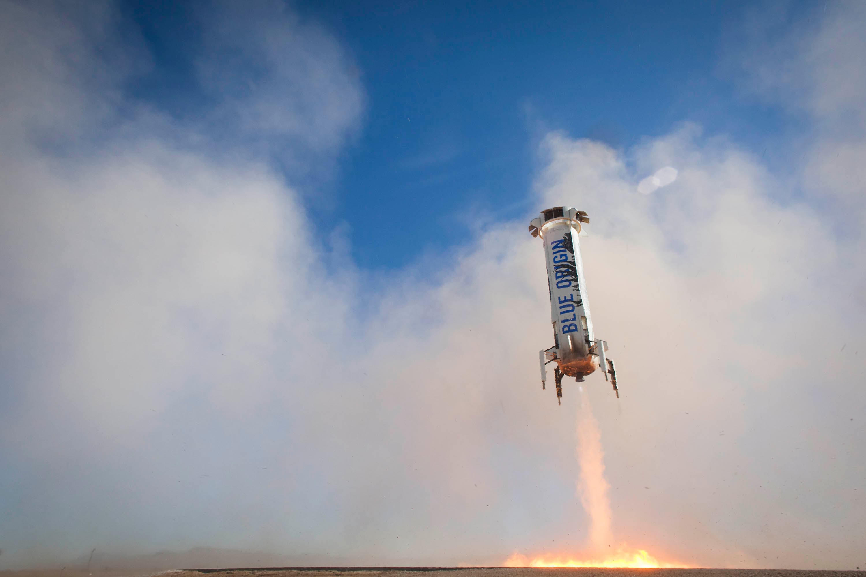 The booster expels pale fiery exhaust and kicks up dust as it descends through the clouds towards the pad.