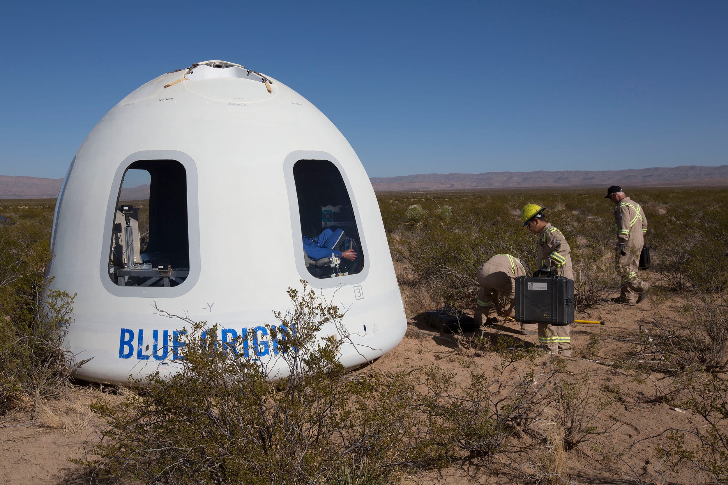 Three Blue Origin employees approach the crew capsule surrounded by desert shrubery. An instrumented test dummy can be seen through the capsule's right window. Mountains rise in the distance beneath a blue sky.