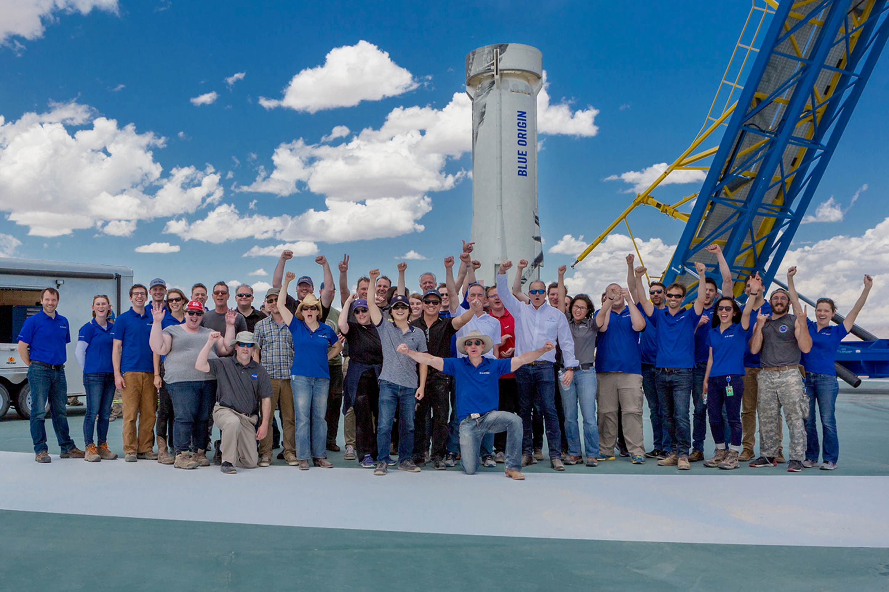 Jeff kneels in the front center of a large group of people wearing a cowboy hat. The group extends their arms in celebration in front of the landed booster in the background.