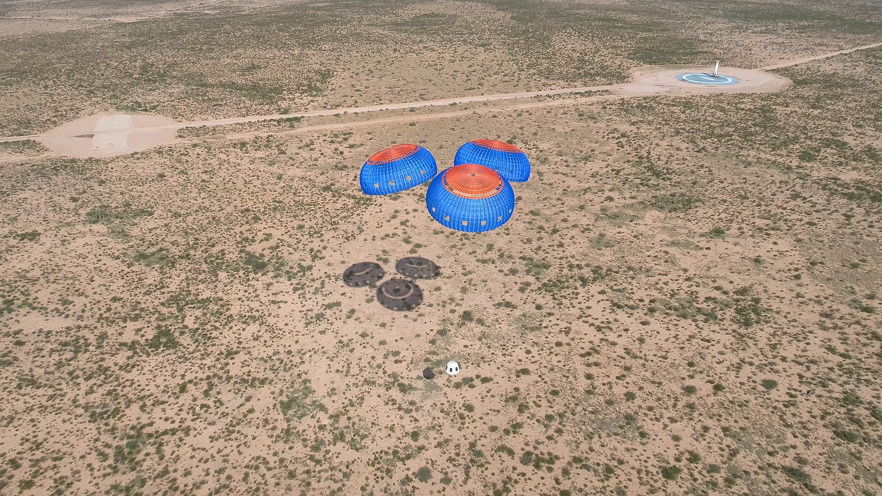 An aerial view of the crew capsule descending under three fully deployed parachutes, which create a shadow on the desert floor. The landed booster on the pad appears as a speck in the far right of the photo; the desert fills the frame.