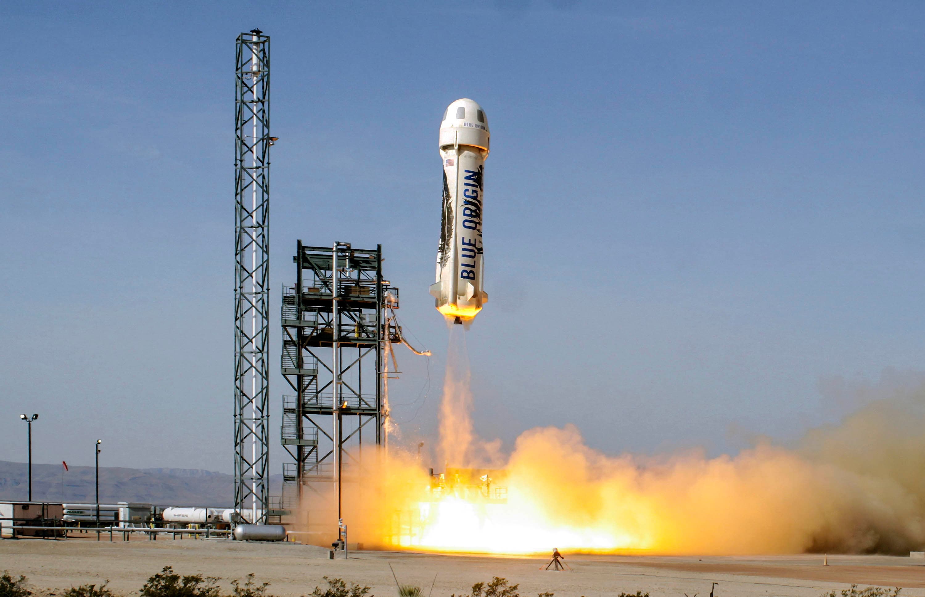 New Shepard moments after liftoff, leaving a cloud of amber fire and white smoke in its wake as the vehicle clears the launch tower and soars towards a bright blue sky.