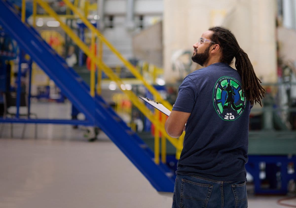 A man with a clipboard on the manufacturing floor wearing a t-shirt from the NS-20 launch