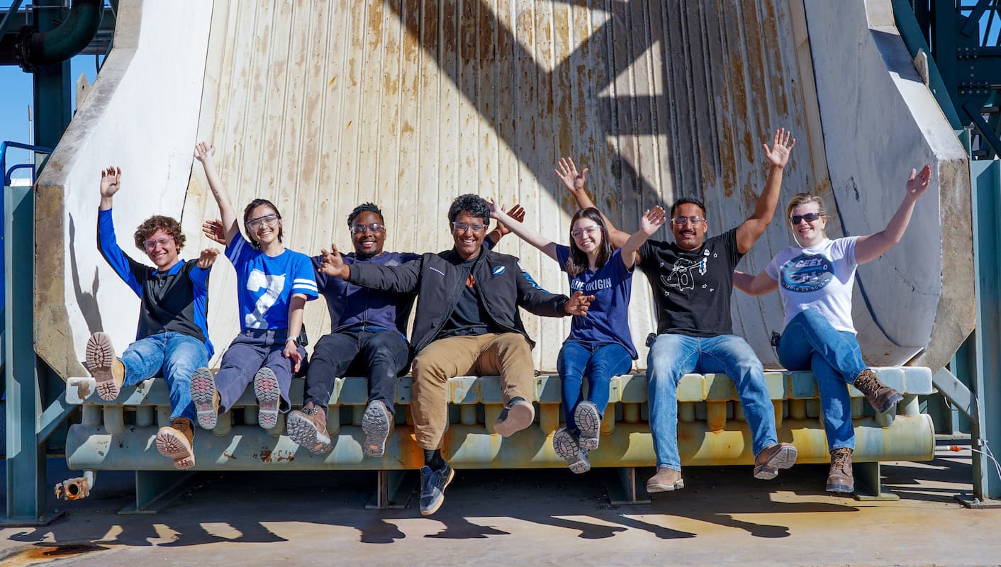 A group of interns in Blue Origin attire sitting with their arms in the air expressing excitement