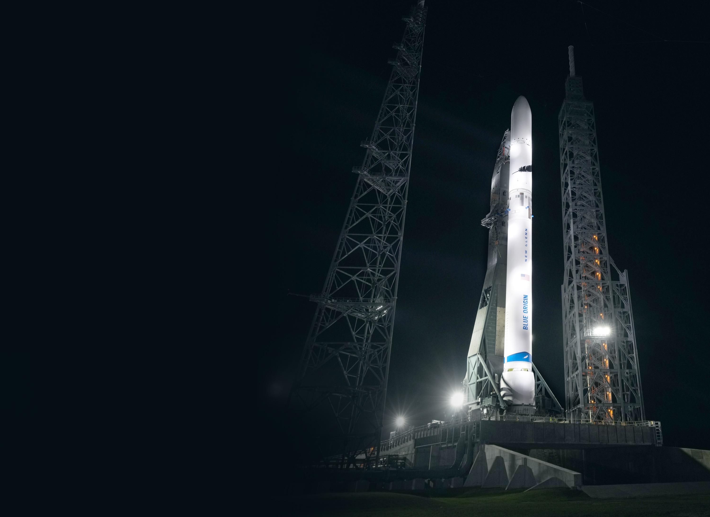 A tall, white rocket lit by spotlights stands upright against the dark night sky.