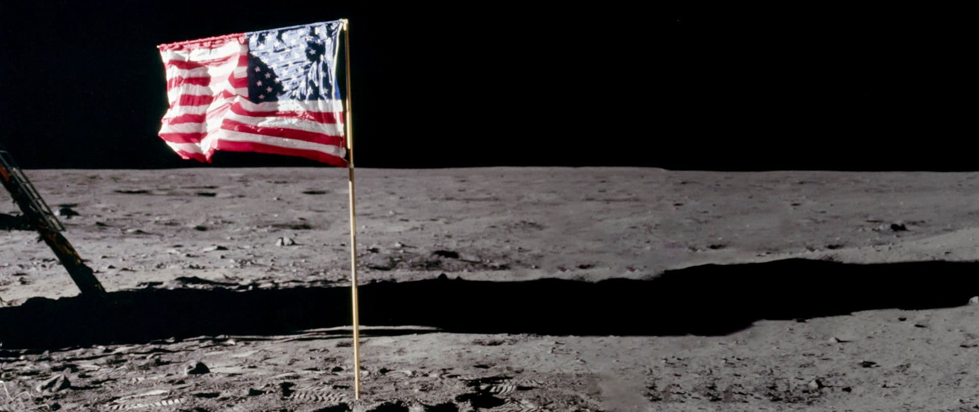 The American flag flies from a gold pole planted on the Moon's surface with visible footprints on the ground and the shadow of a lunar lander in the background.