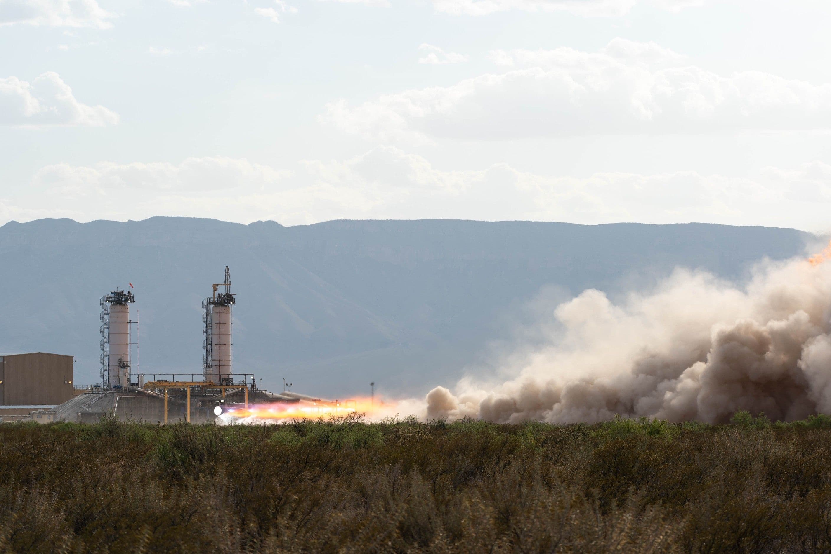 A BE-4 is hotfired on the test stand during the day, expelling fiery exhaust that transforms into horizontal white plumes of smoke. Desert scrub brush appears in the foreground and mountains appear in the distance against a hazy sky.