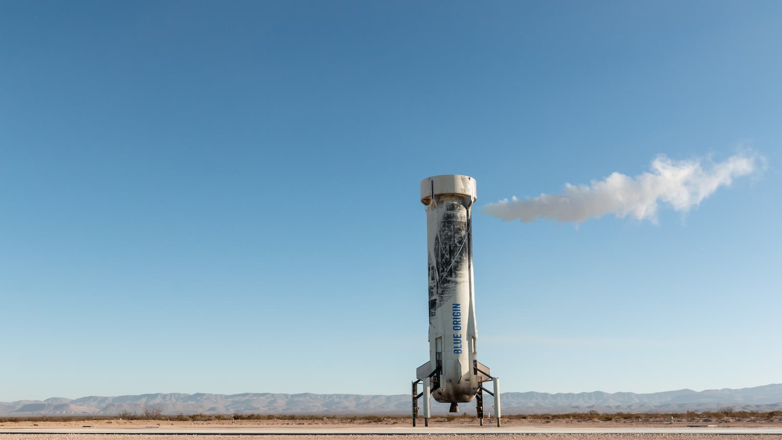 A wisp of white exhaust horizontally expels from the top of the landed booster; mountains and clear blue sky appear in the distance.