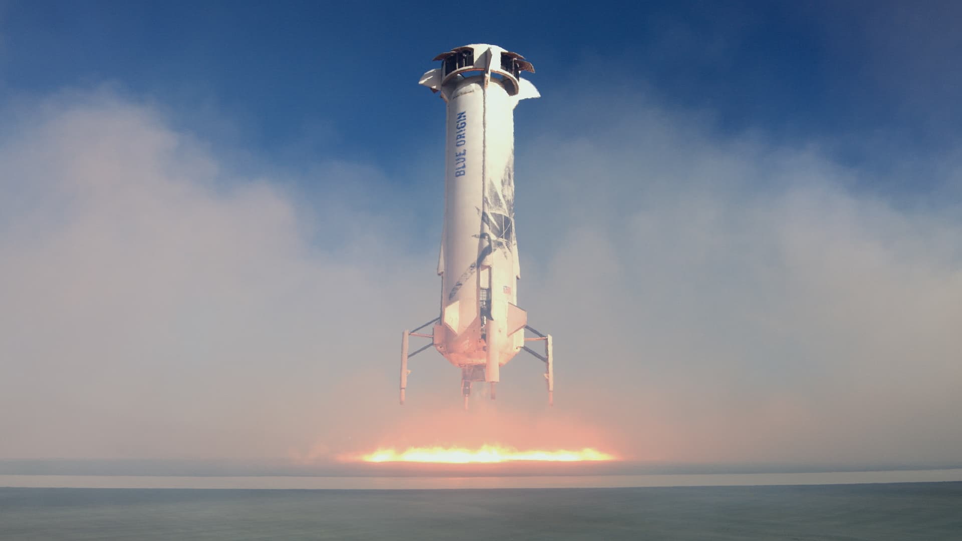 The booster hovers above the pad, expelling an orange circle of fiery exhaust. Pale white smoke fills the frame against a blue sky.