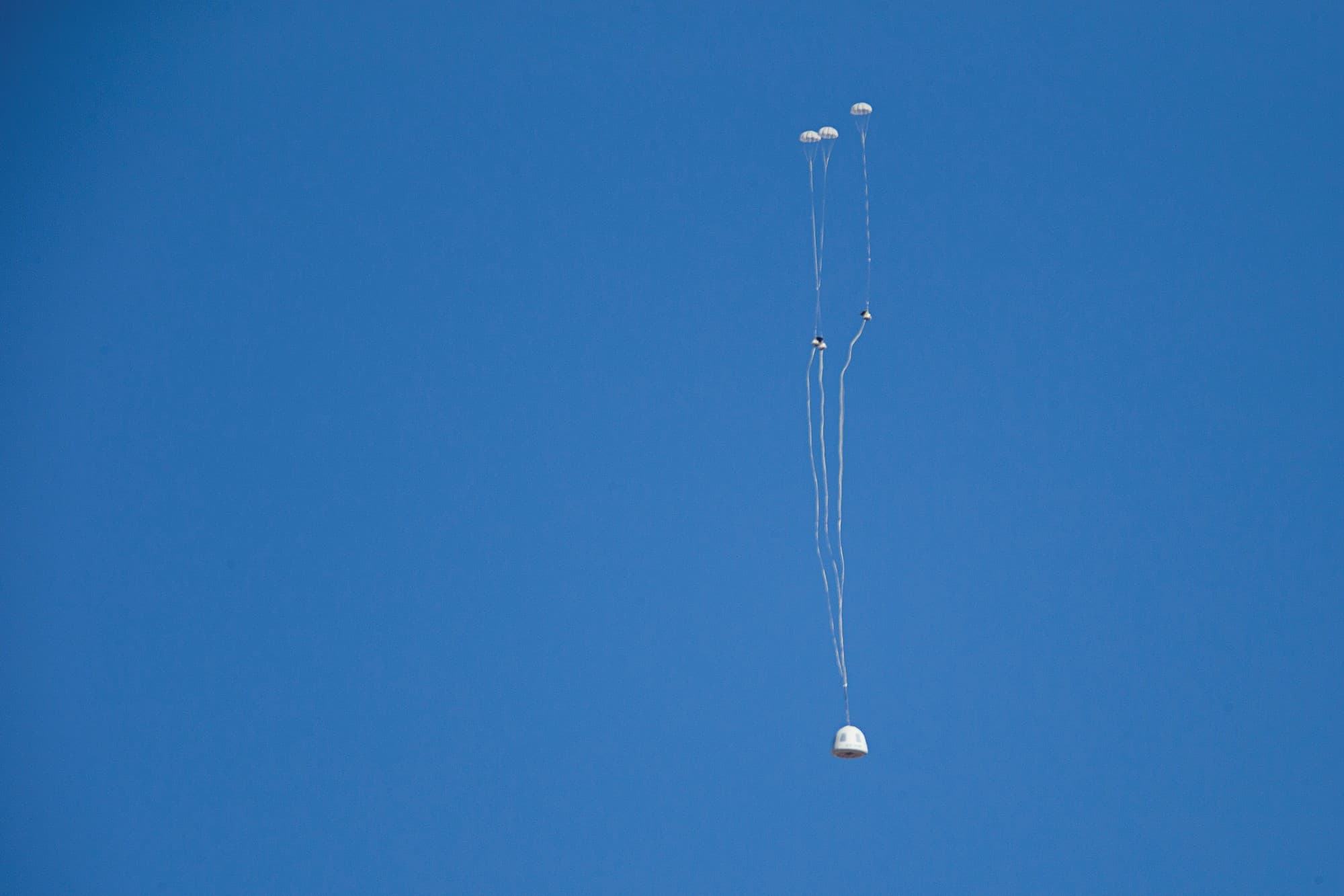 The crew capsule descends under parachutes against a bright blue sky.