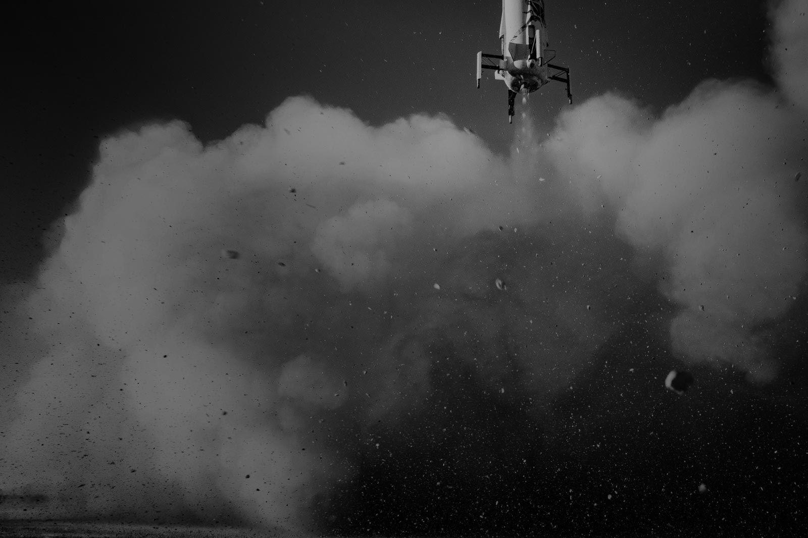 A black-and-white photo of the bottom of the booster, including the landing legs. Thick clouds of dust, smoke, and small rocks fill the frame.