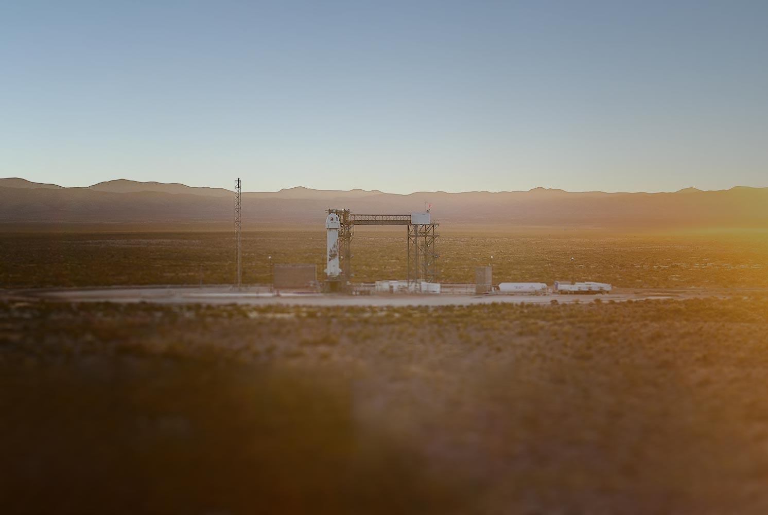 New Shepard is perched on the launch tower in the distance. The sun rises above the desert surrounding the tower.