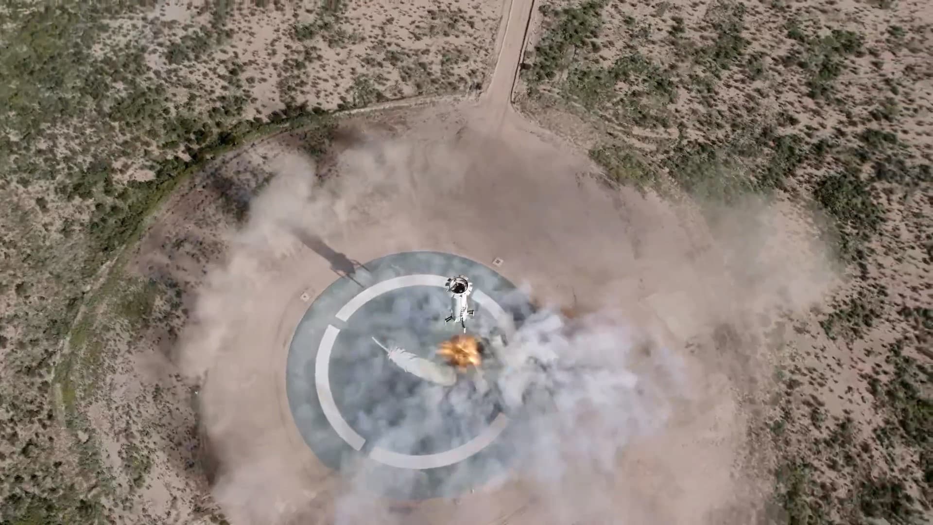 An aerial view of the New Shepard booster descending onto the landing pad with fiery exhaust expelling below and scrub brush seen in the distance.