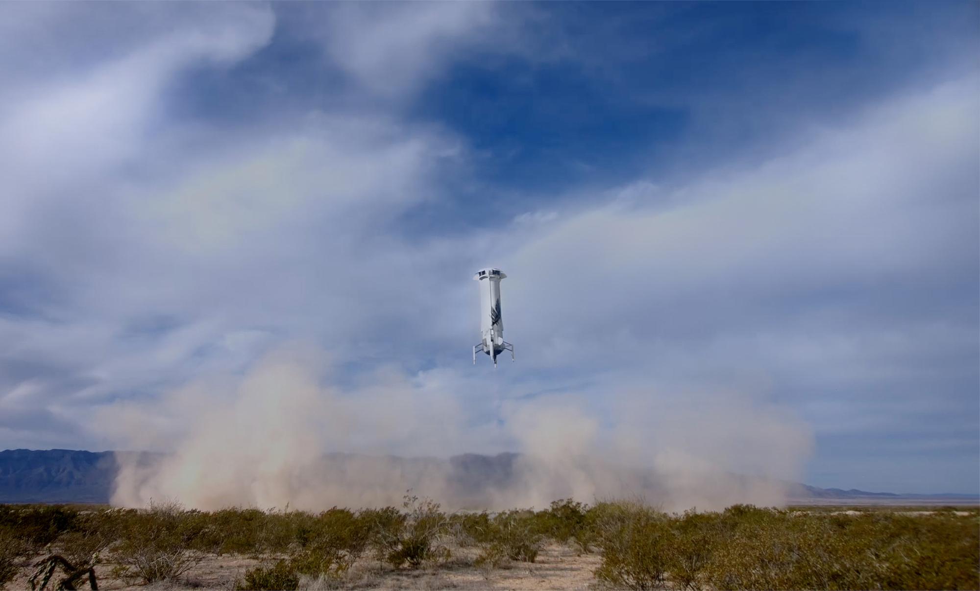 New Shepard’s booster kicks up dust prior to landing on the pad in the West Texas desert. There are clouds in the sky and scrub brush in the foreground.