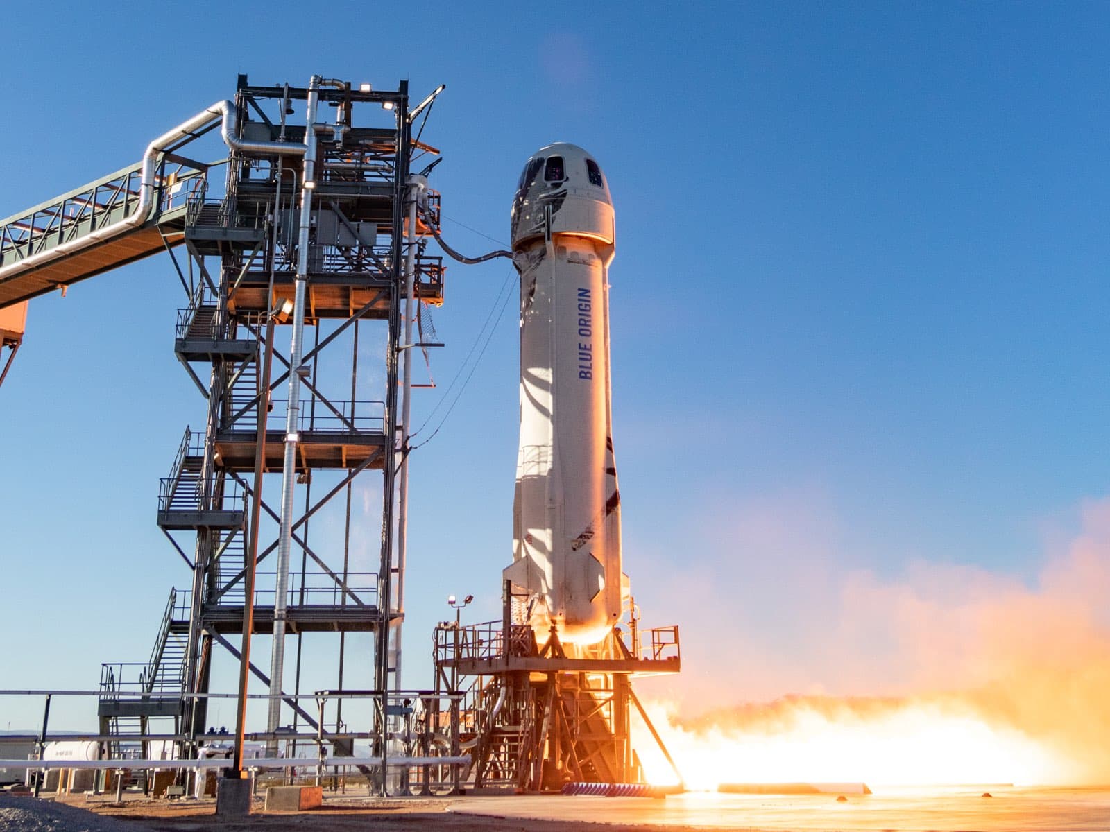 New Shepard next to the launch tower at the moment of ignition. Fiery exhaust expels from the bottom of the booster. Blue sky appears in the background.