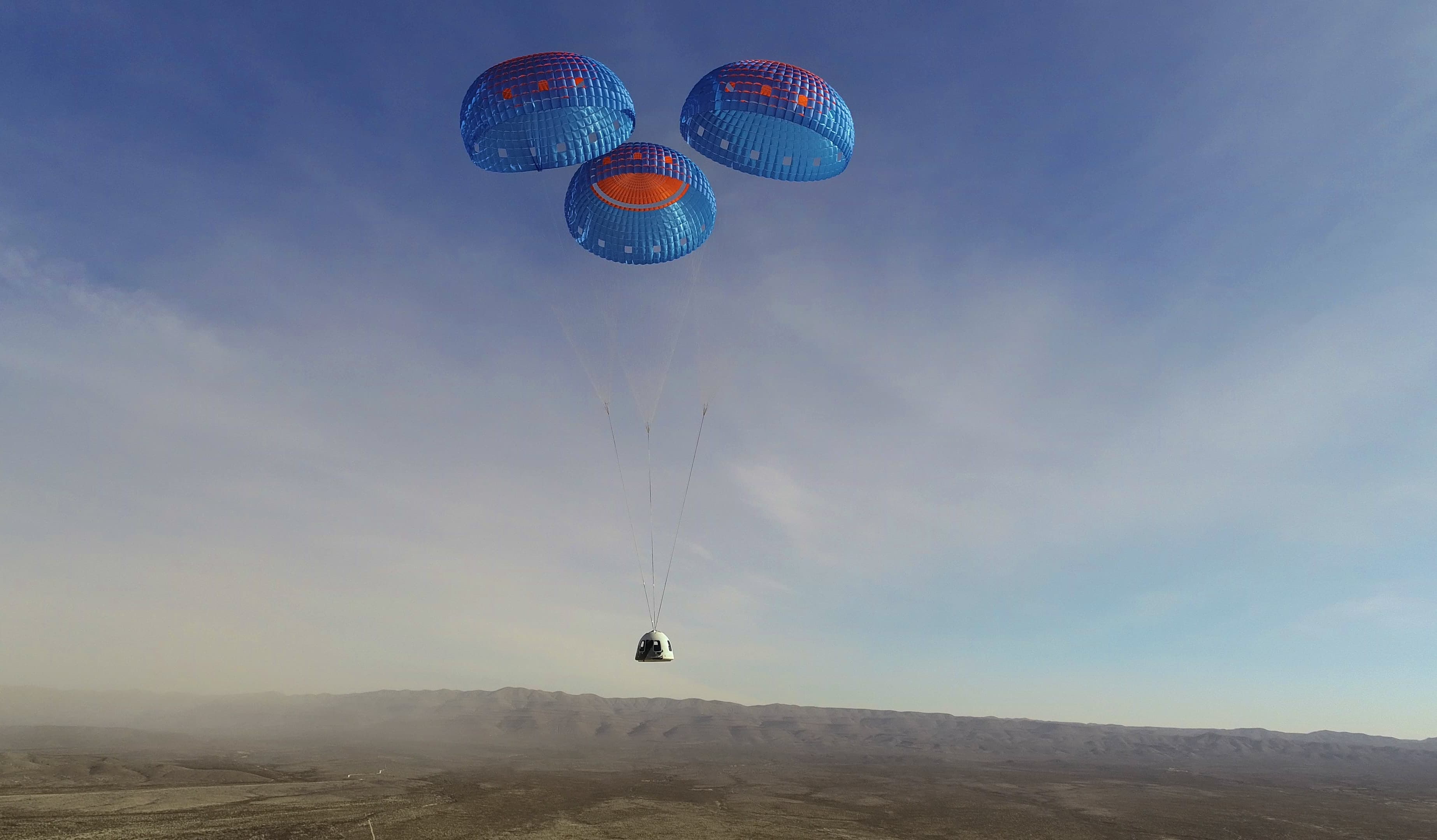 The crew capsule descends under its three main blue and orange main parachutes; the capsule looks tiny against the vastness of the desert and mountains in the background.