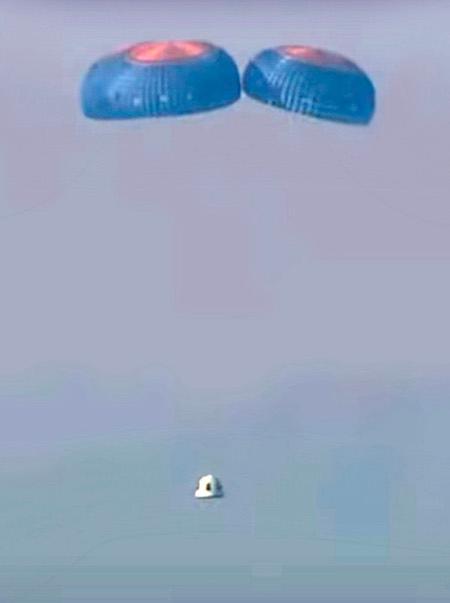 The white crew capsule descends under its three main blue and orange parachutes.