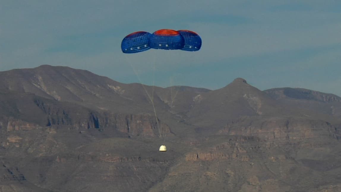 The dome-shaped crew capsule floats towards Earth under its three main blue and orange parachutes. Mountains can be seen in the distance.