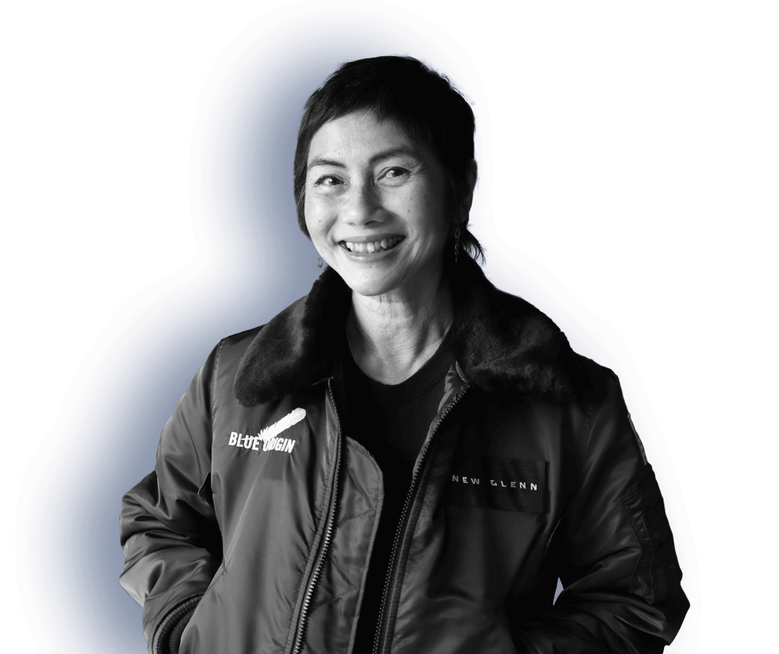 A woman with short hair smiles, standing with her hands in the pockets of a New Glenn branded jacket with a fuzzy collar.